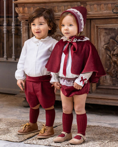 Two children in matching outfits with a maroon cape and white shirts, standing in front of a wooden fireplace.