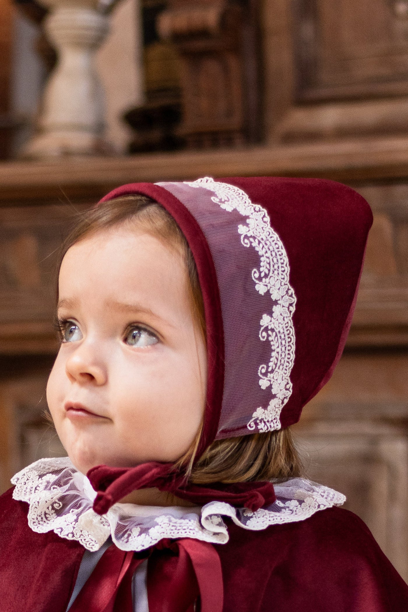 Child wearing a red costume with a hood and white lace details in a rustic setting.