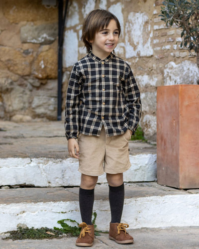 Child wearing a plaid shirt, beige shorts, and brown shoes standing in front of a stone wall.