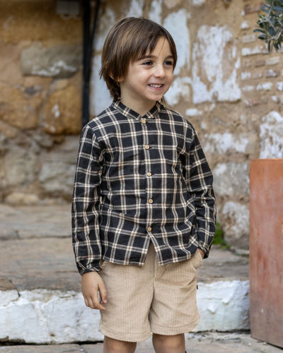 Child wearing a plaid shirt and shorts standing in front of a stone wall.