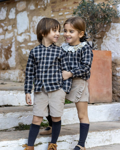 Two children wearing matching plaid shirts and shorts standing on a stone path.