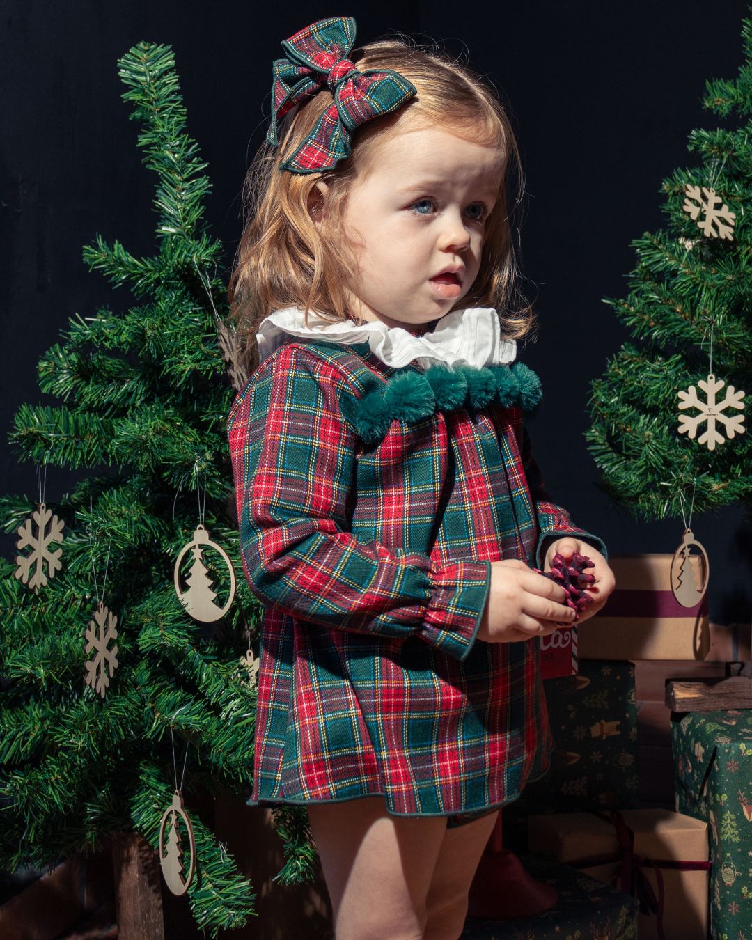 Young girl in a plaid dress with a bow, standing in front of Christmas trees and decorations.