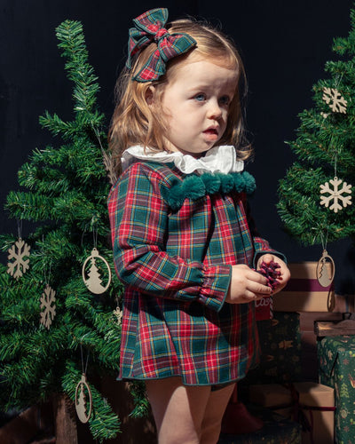 Young girl in a plaid dress with a bow, standing in front of Christmas trees and decorations.